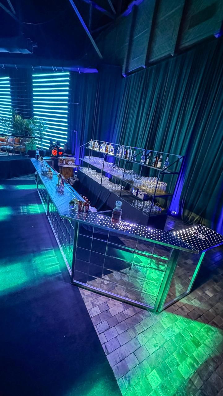 Barra espejada con iluminación verde y azul, estantería de botellas y copas de fondo