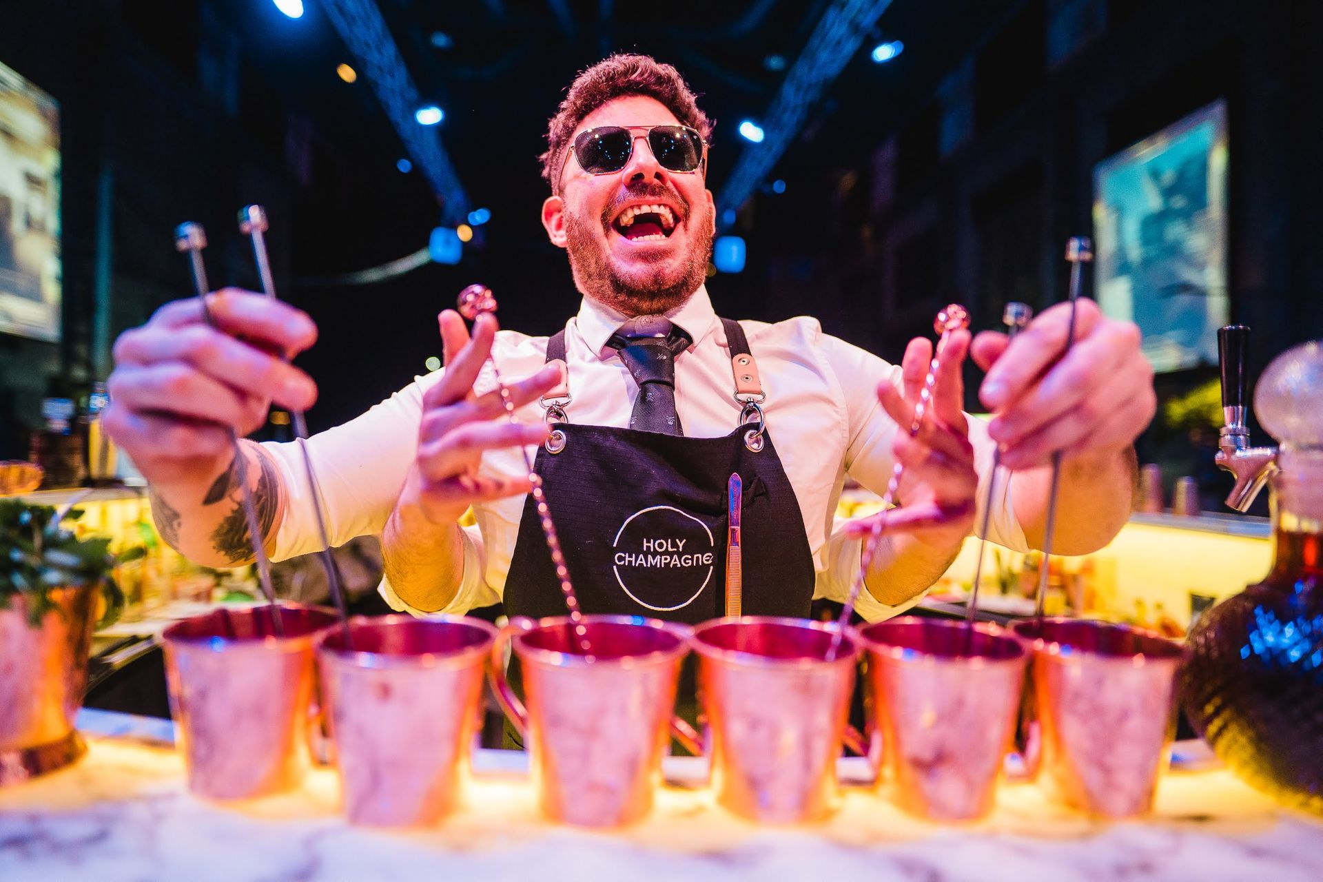 Bartender de Holy Champagne sonriendo y presentando una fila de vasos de cobre listos para servir