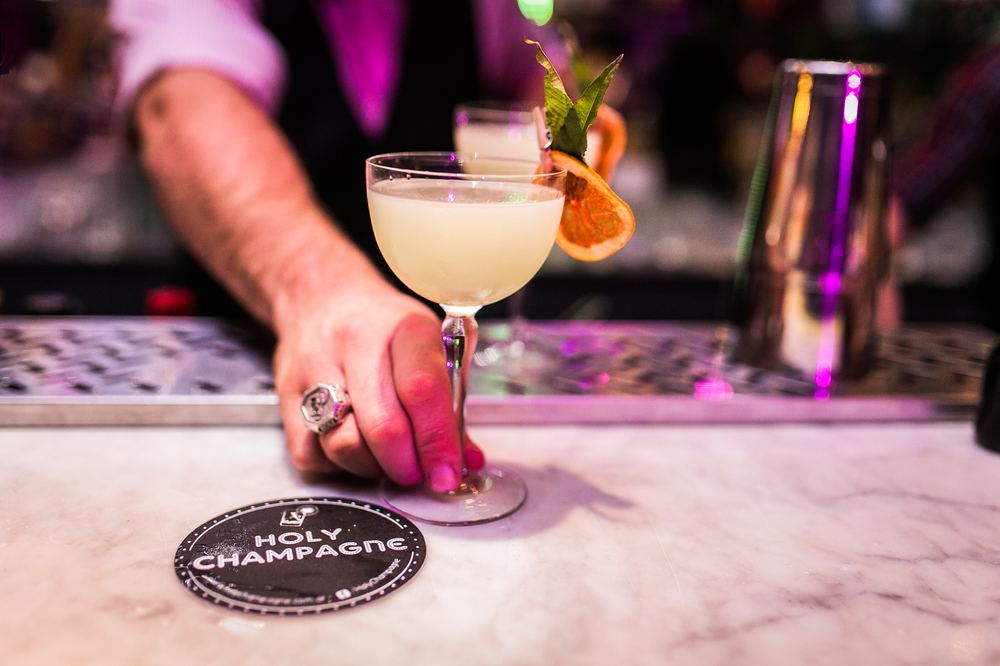 Bartender sirviendo un cocktail en copa sobre la barra, con posavasos de la marca Holy Champagne