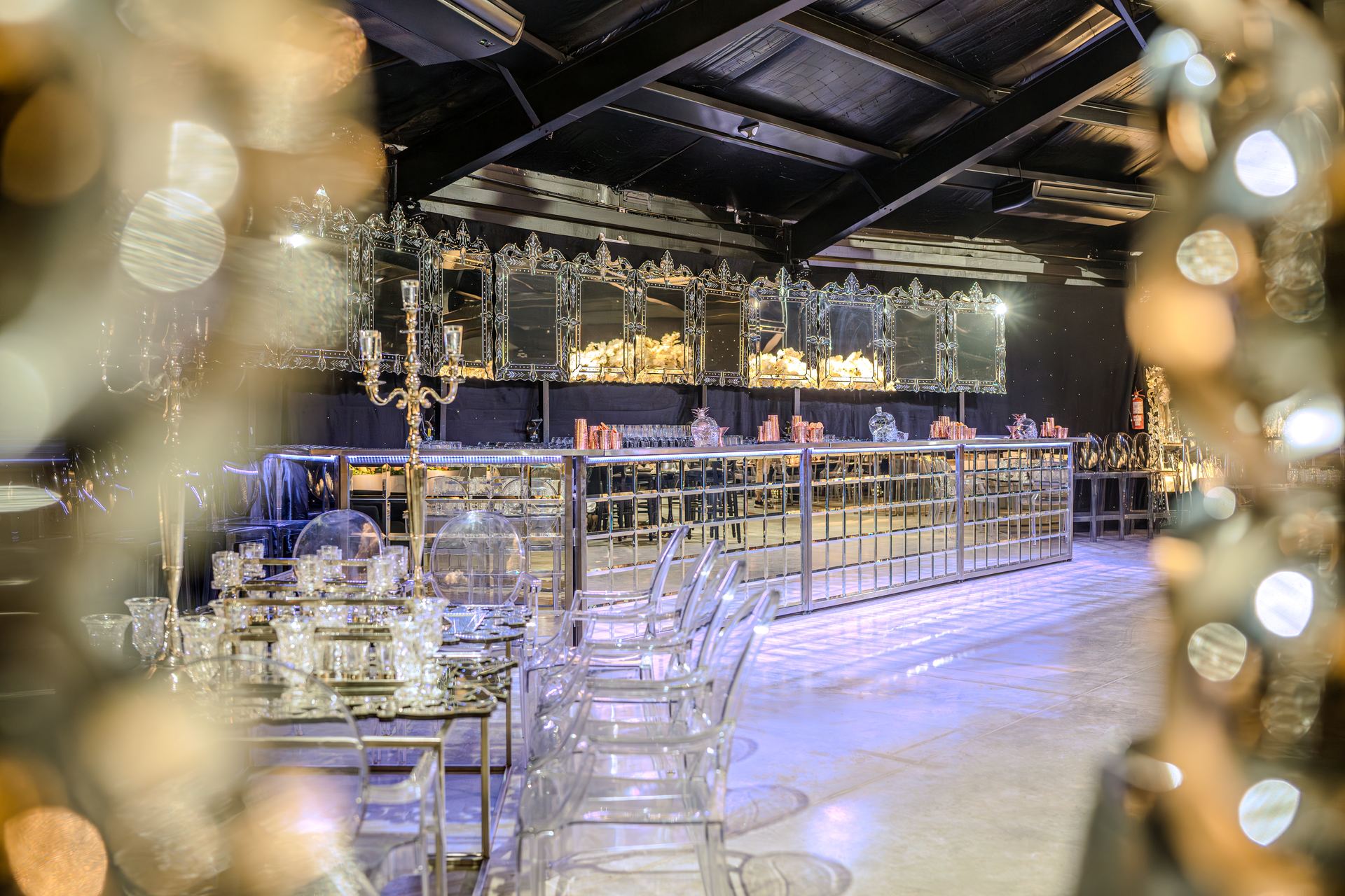 Barra móvil Holy Champagne espejada bajo espejos venecianos, con candelabro y sillas transparentes en un salón de evento