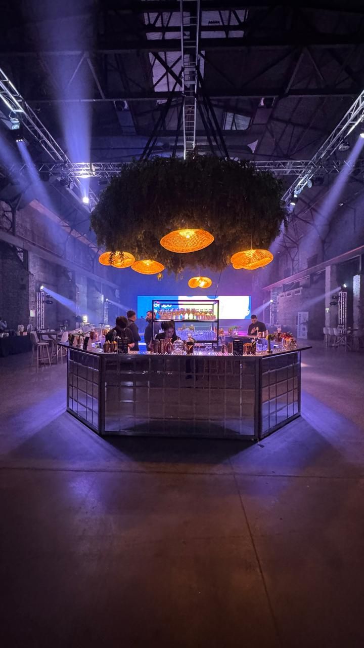 Isla Bar hexagonal espejada en el centro de un salón industrial, con luces violetas y follaje colgante