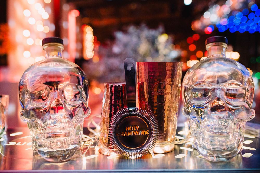 Coctelera de cobre con branding Holy Champagne entre botellas con forma de calavera