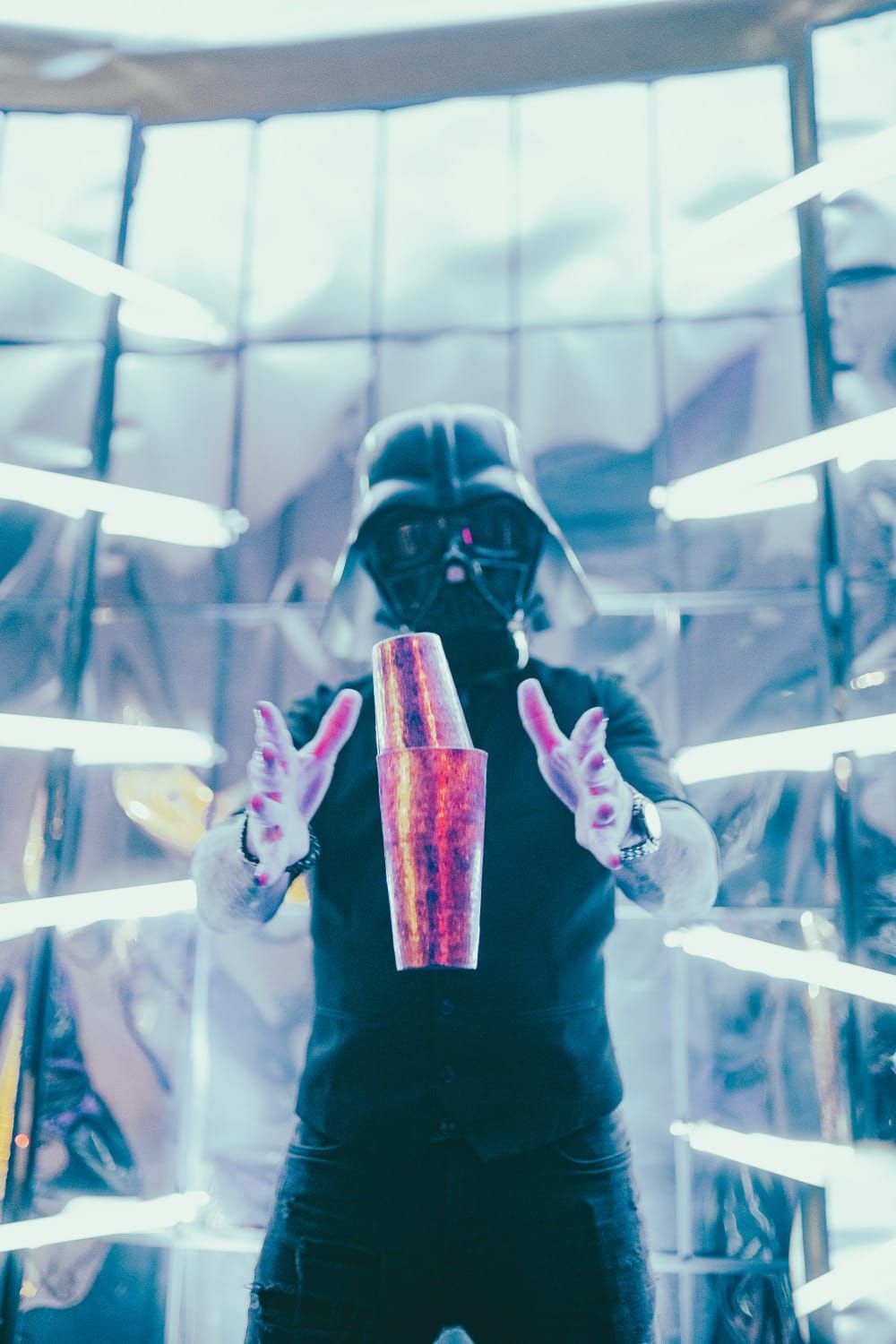 Bartender enmascarado lanzando una coctelera al aire frente a una barra espejada con luces